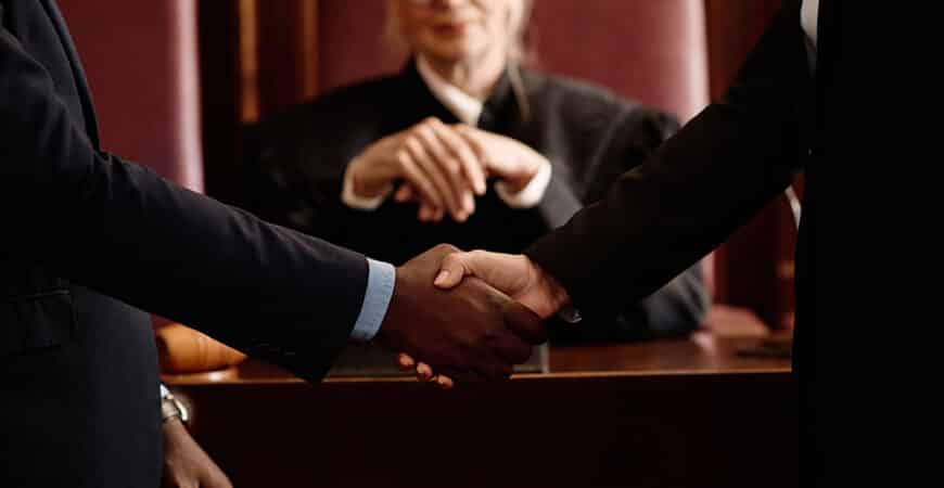 Close-up of two opposite sides of court case shaking hands after making agreement while standing in front of camera against female judge